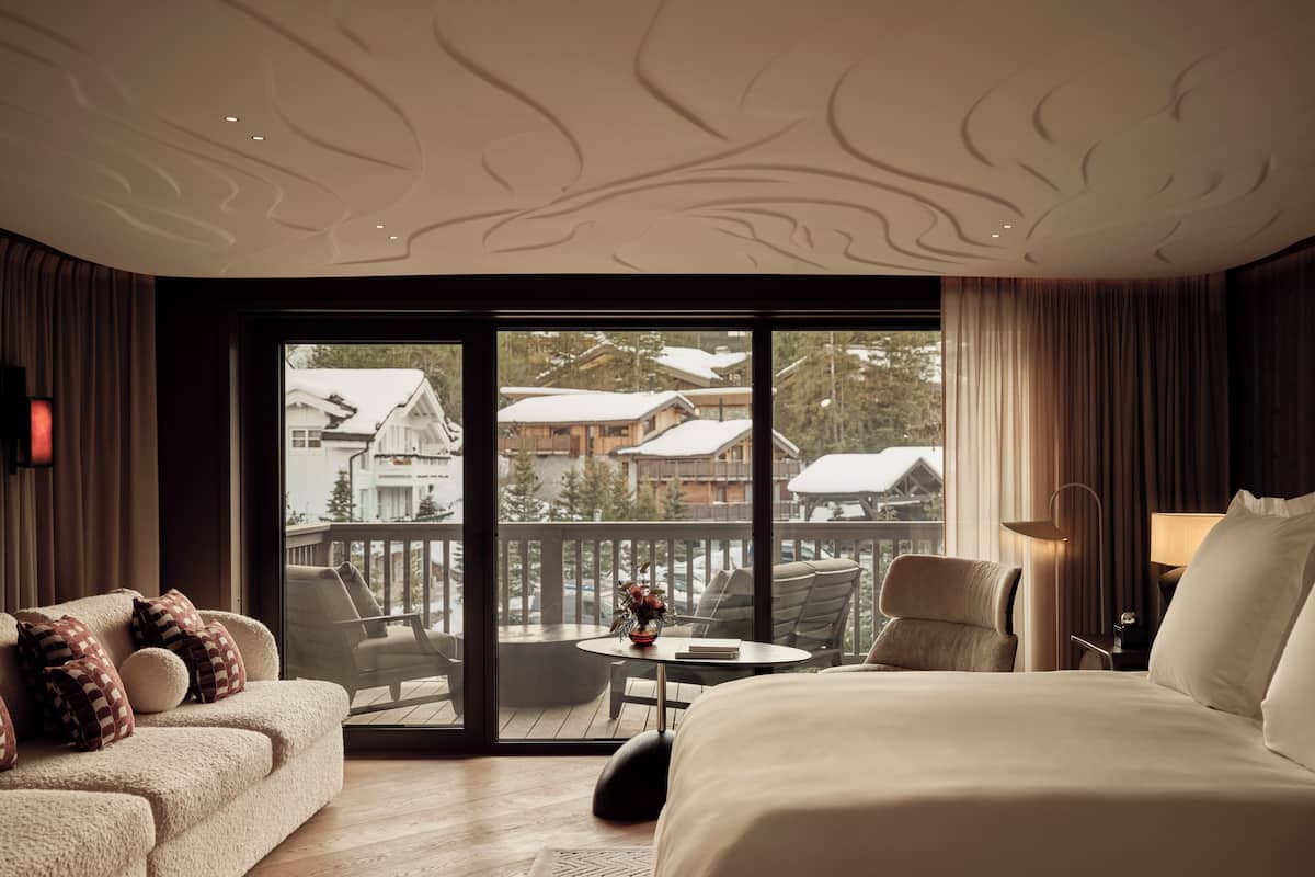 A hotel room with a large bed, a sofa, a dining table with chairs, and a view of snow-covered buildings through large windows.