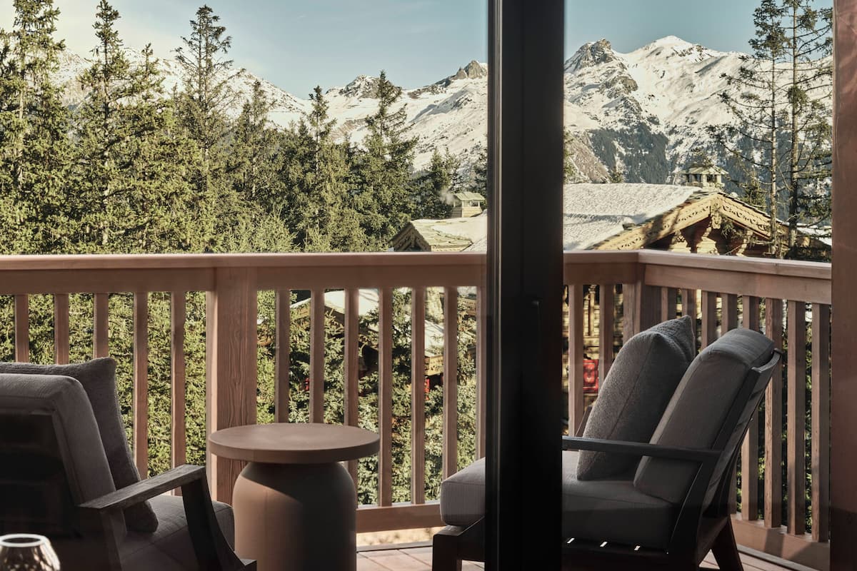 A wooden deck with a table and chairs, overlooking a mountainous landscape with snow-capped peaks and dense forest.