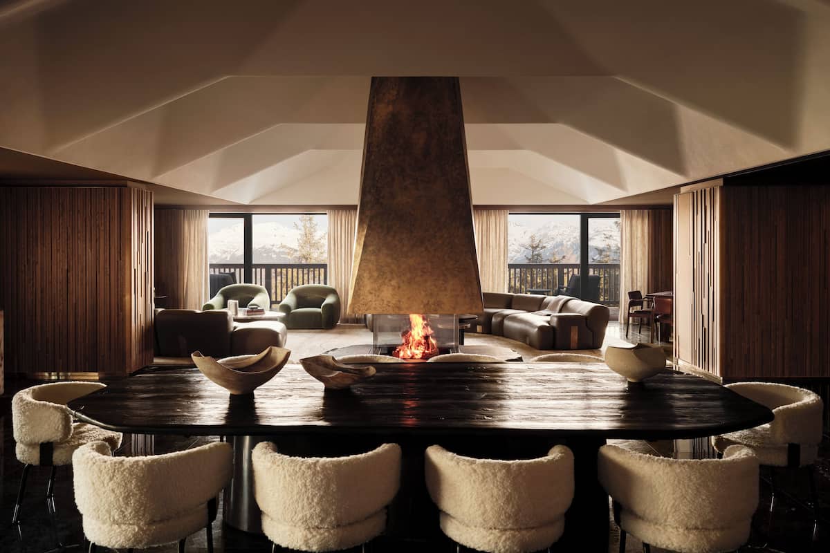 A spacious living room with a large round table, plush chairs, a fireplace, and a view of snow-capped mountains through large windows.