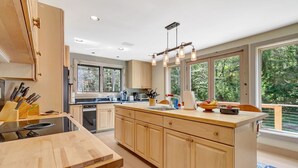 A modern kitchen with wooden cabinets, a central island, and large windows offering a view of the outdoors.