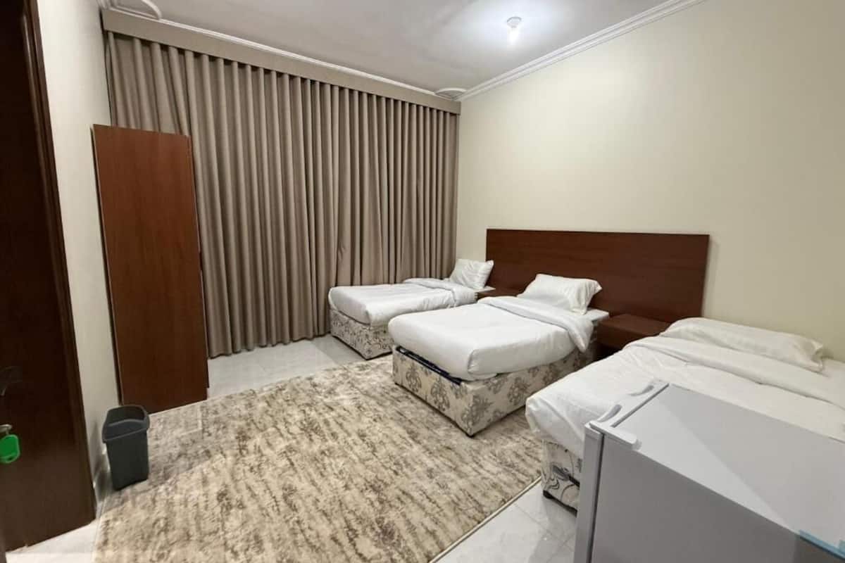 A hotel room with two single beds, a large window with curtains, a wooden wardrobe, and a small trash bin.