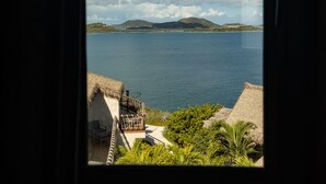 A view from a window overlooking a calm body of water, with a thatched-roof structure and greenery in the foreground. - The Dream | Private 360° View Suite in Buck Island (Buck Island)