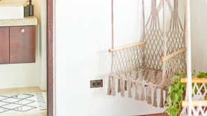 A room with a wooden floor, a round woven rug, a macrame hanging chair, a white wall, and a wooden cabinet with a mirror.