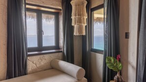 Een kamer met een bank, een hangende macramé lamp, een potplant en uitzicht op de zee door grote ramen.