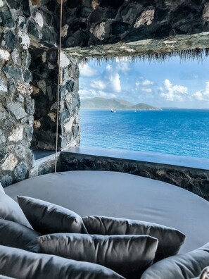 A stone-walled room with a view of the sea and a bed with grey cushions. - Top‑Level Sunrise Oceanview Suite, Private Balcony (Buck Island)