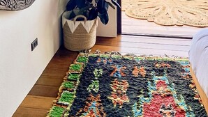 A room with a wooden floor, a colorful rug, a plant in a woven basket, and a view of the sea through a balcony with a rope railing.