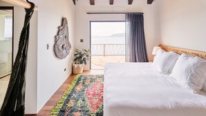 A bedroom with a bed, a colorful rug, a plant, and a view of the beach.
