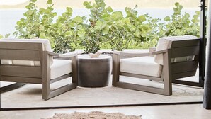Un rincón con asientos para dos personas, con dos sillas y una mesa pequeña, rodeado de plantas en macetas, con vistas a una playa y montañas.