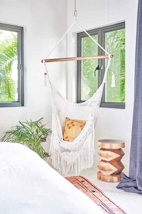 A hammock chair in a room with a bed, a wooden stool, and a plant.
