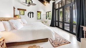 Une chambre spacieuse avec un grand lit, un ventilateur de plafond et une vue sur l’extérieur grâce à de grandes fenêtres.