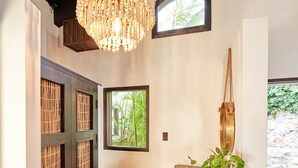 Un couloir avec une porte en bois, un tapis tissé, un banc en bois agrémenté d’une plante, un luminaire suspendu et une fenêtre donnant sur un paysage verdoyant.