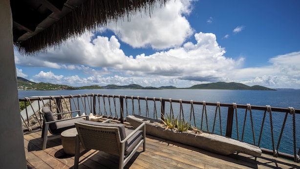Una terraza de madera con mobiliario exterior, techo de paja y vistas al mar y colinas distantes.