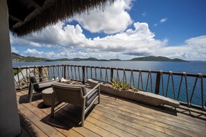 A wooden deck with outdoor furniture, a thatched roof, and a view of the sea and distant hills. - Ground‑Level Sunrise Oceanview Suite in BVI (Buck Island)