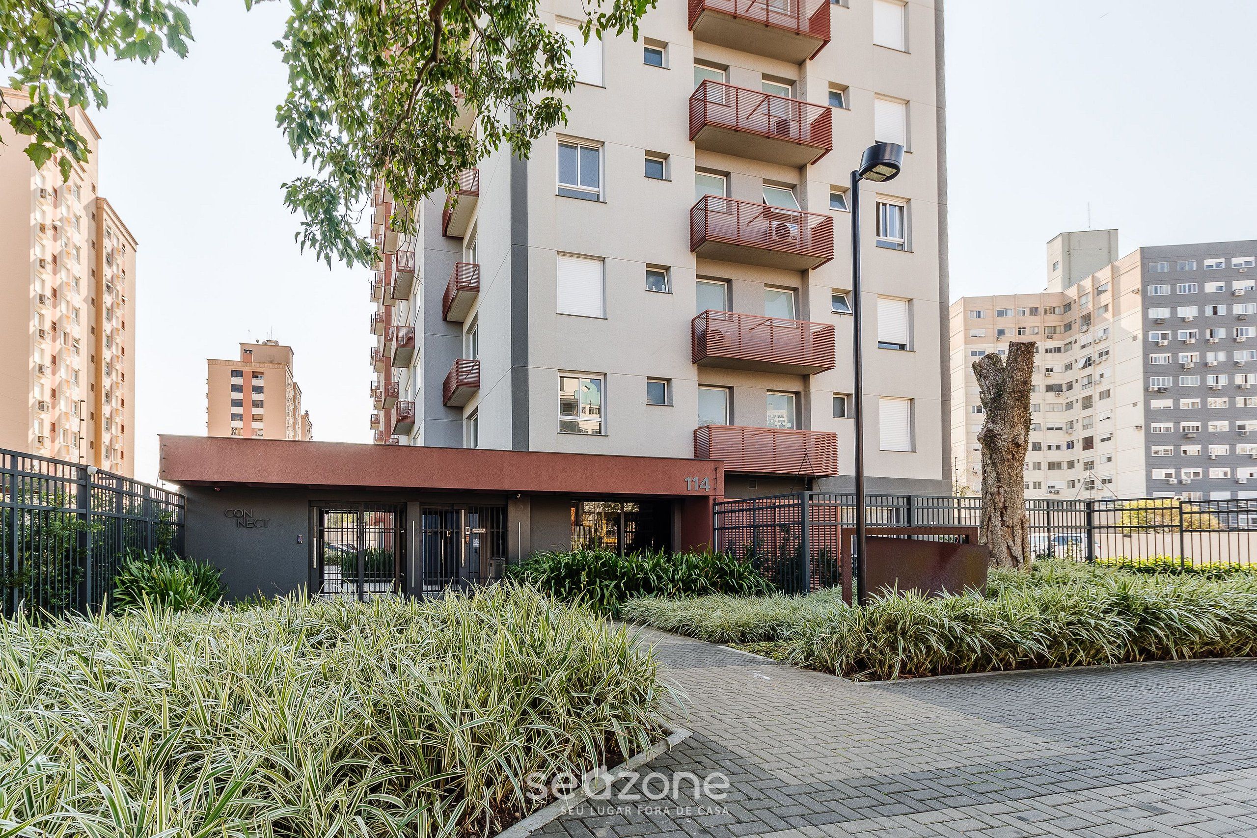 Un moderno edificio de apartamentos con balcones, entrada protegida por un portón y un área ajardinada con plantas.