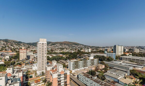 A cityscape with a prominent high-rise building, a mix of residential and commercial buildings, and a hilly background. - Studio Completo ao Lado da PUCRS c/ Garag CON1506 (Porto Alegre)
