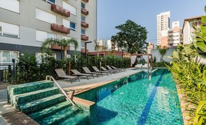 A swimming pool with lounge chairs and a wooden deck, surrounded by greenery and modern buildings. - Studio Completo ao Lado da PUCRS c/ Garag CON1506 (Porto Alegre)