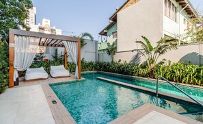 Ein Pool mit Pergola und Liegestühlen, umgeben von Grünpflanzen und einem Gebäude.