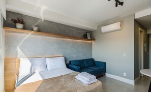 A modern bedroom with a bed, a blue sofa, a wooden shelf with decorative items, and an air conditioning unit. - Studio Completo ao Lado da PUCRS c/ Garag CON1506 (Porto Alegre)
