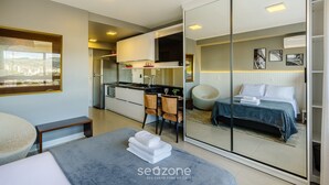 A modern hotel room with a large bed, a seating area with a bench, a small kitchen, and a city view. - Studio in Building With Pool Con1206 (Porto Alegre)