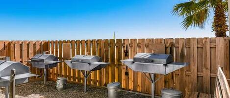 An outdoor barbecue area with three grills, a wooden fence, and a palm tree.