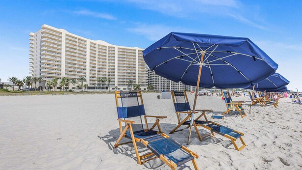 Ein Strand mit Liegestühlen und Sonnenschirmen, ein großes Hotelgebäude im Hintergrund und Palmen.