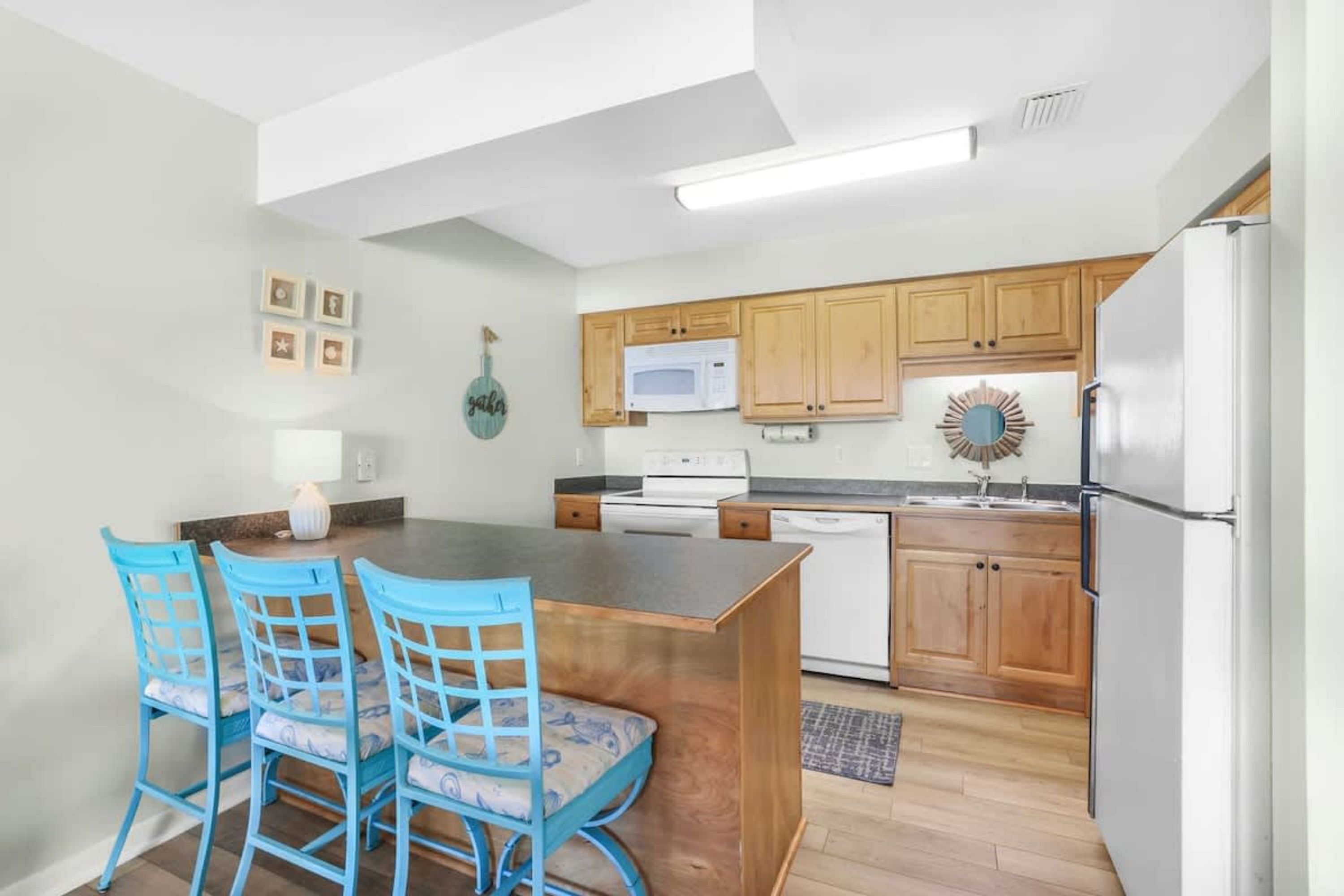 A modern kitchen with wooden cabinets, a central island, blue chairs, a microwave, and a refrigerator.