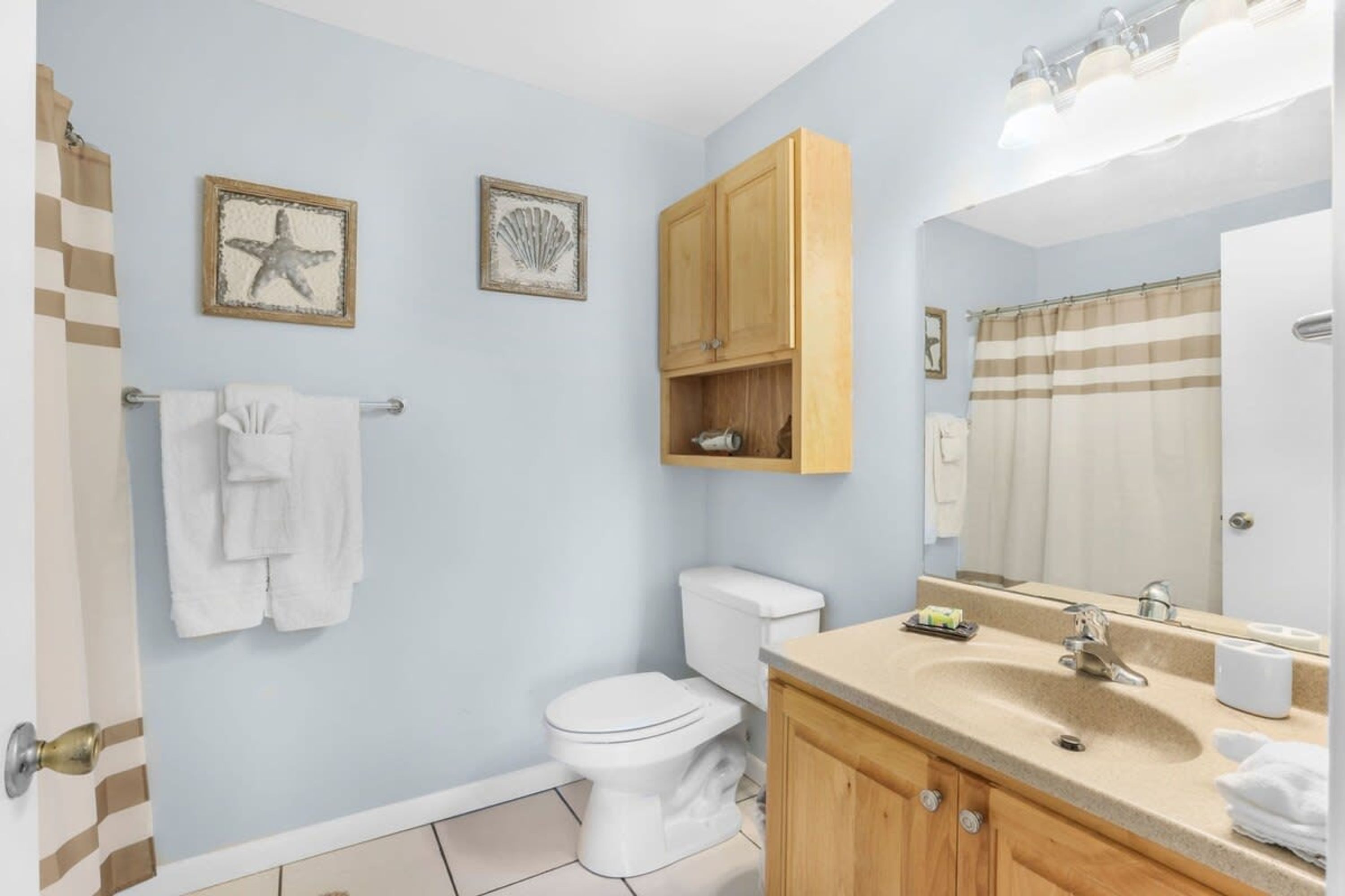 Un bagno con il water, un lavandino e uno specchio. In evidenza due quadri appesi al muro, un porta asciugamani e un mobiletto in legno sopra il lavandino.