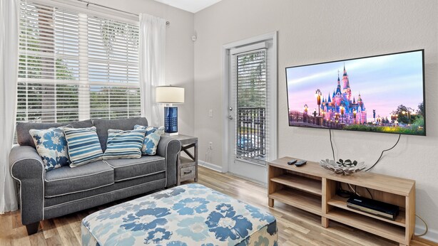 Un salon avec un canapé gris, un pouf bleu à motifs floraux, un téléviseur diffusant une image de château et un meuble TV en bois.