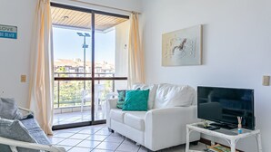Ein Wohnzimmer mit einem weißen Sofa, einem Fernseher auf einem kleinen Tisch und Blick ins Freie durch ein großes Fenster.