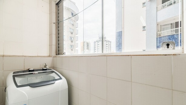 A white washing machine in a room with white tiled walls and a window overlooking a cityscape. - Apt 400m fr Beach Close to Rest. n Markets Cev1106 (Balneário Camboriú)