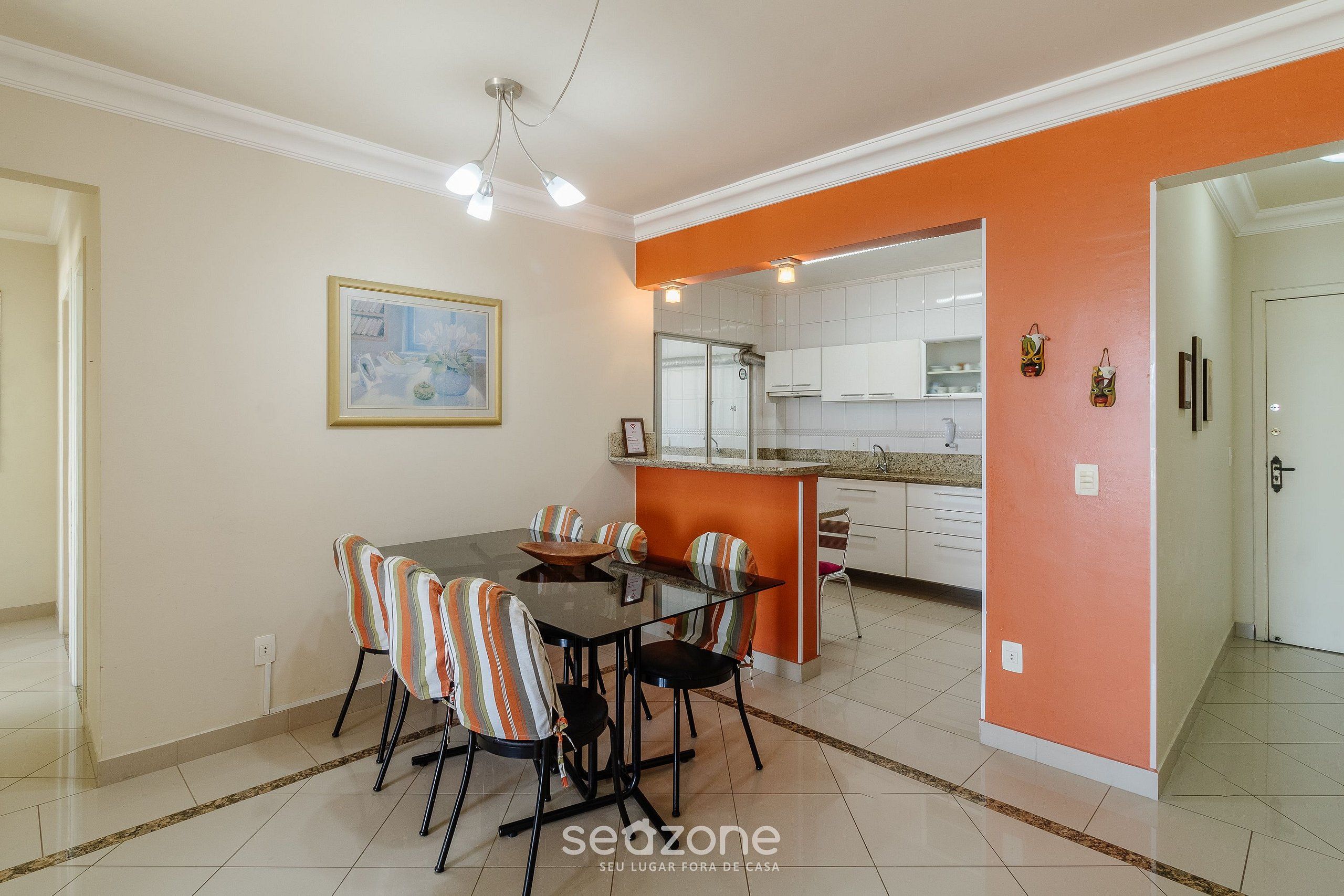 Un comedor con mesa de vidrio y sillas a rayas, una pared de acento naranja y una cocina al fondo.