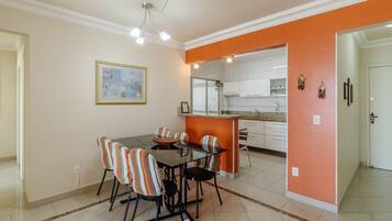 A dining area with a glass table and striped chairs, an orange accent wall, and a kitchen in the background.