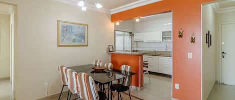 A dining area with a glass table and striped chairs, an orange accent wall, and a kitchen in the background.
