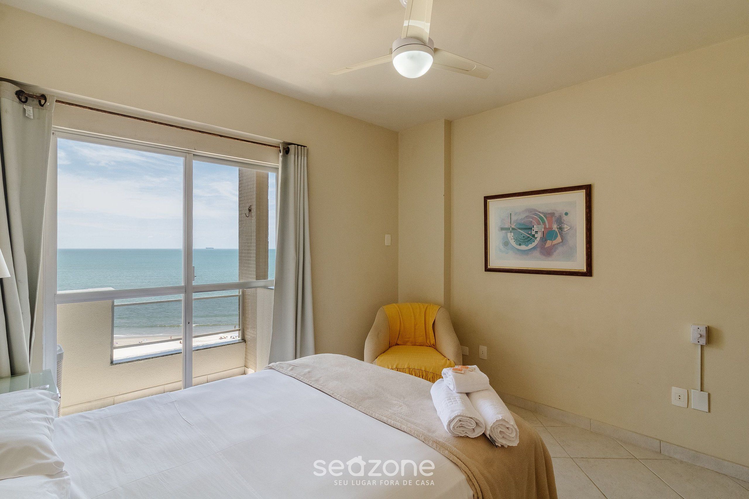 Ein Hotelzimmer mit großem Fenster und Meerblick, einem Bett mit ordentlich gefalteten Handtüchern, einem Sessel mit gelber Sitzkissen und einem Deckenventilator.