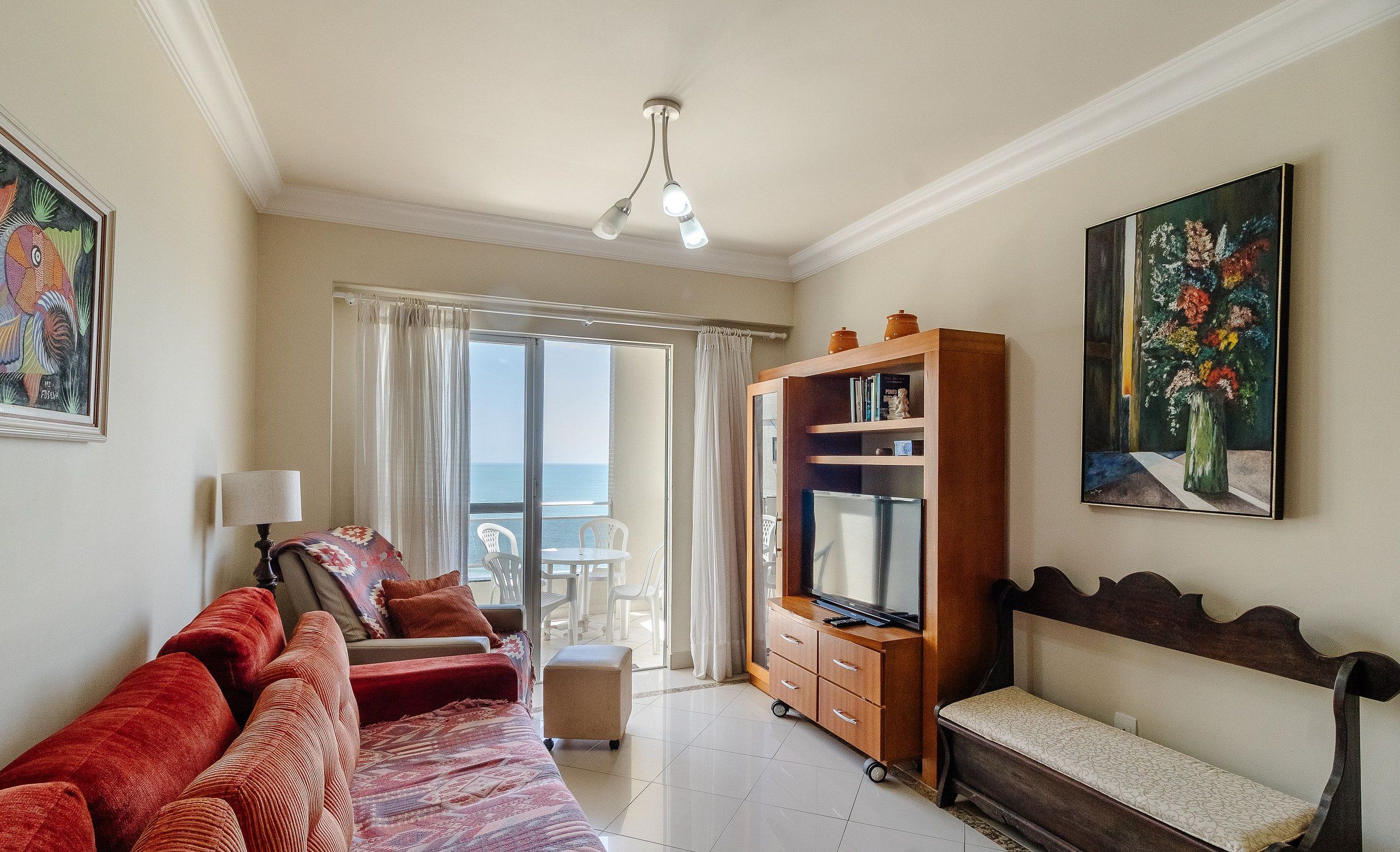 A spacious living room with a sofa, a television, and a bench. There is a balcony with a view of the sea.