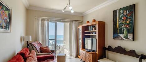 A spacious living room with a sofa, a television, and a bench. There is a balcony with a view of the sea.