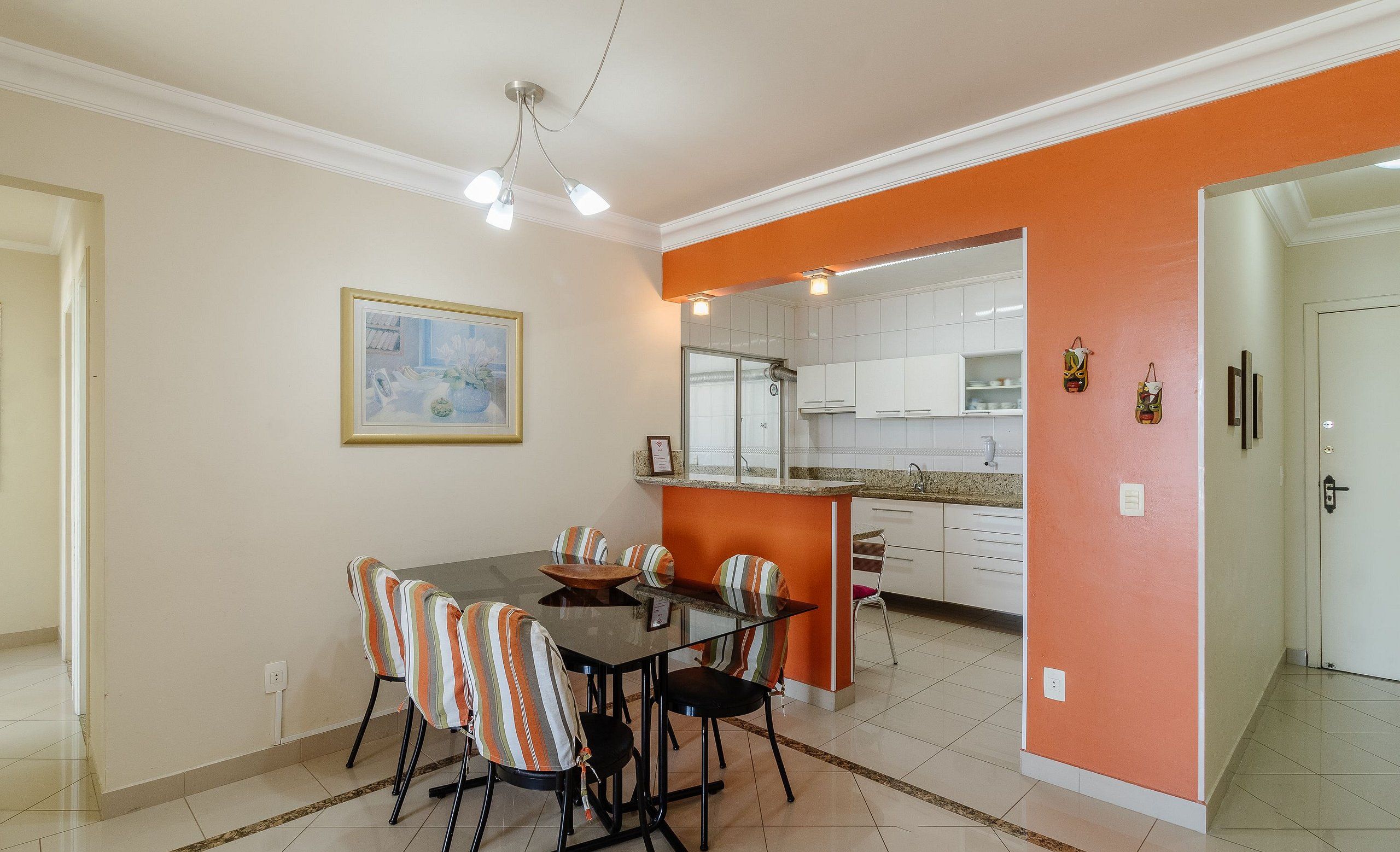 A dining area with a glass table and striped chairs, an orange accent wall, and a kitchen in the background.