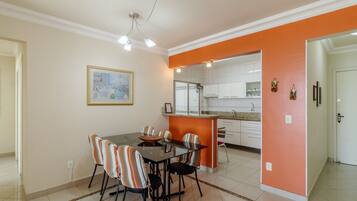A dining area with a glass table and striped chairs, an orange accent wall, and a kitchen in the background.