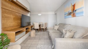 A modern living room with a flat-screen TV, wooden shelves, a beige sofa, and a dining area with chairs and a table.
