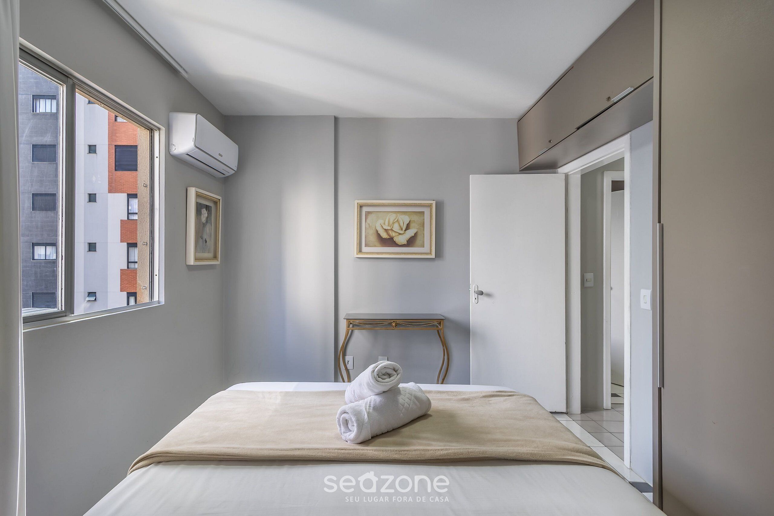 A hotel room with a bed, a framed picture, an air conditioning unit, and a view of buildings outside.