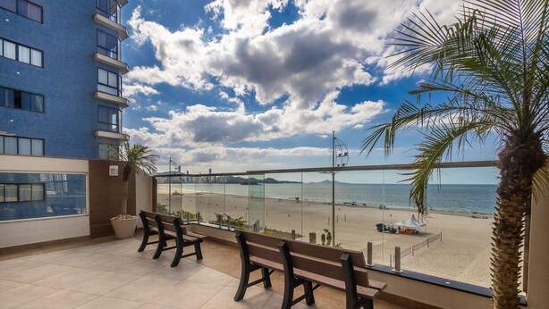 A balcony with a view of a beach, a palm tree, and a building in the background.