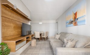 A modern living room with a flat-screen TV, wooden shelves, a beige sofa, and a dining area with chairs and a table. - Beautiful Oceanfront apt Next to Brahma Cet603 (Balneário Camboriú)