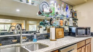 Cocina con encimera de granito, pileta de acero inoxidable y repisa de vidrio sobre la pileta donde se exhiben varias botellas y una camiseta de fútbol.