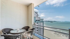 Balcón con sillas de mimbre y una mesa de vidrio con vista a la playa y al paisaje urbano.