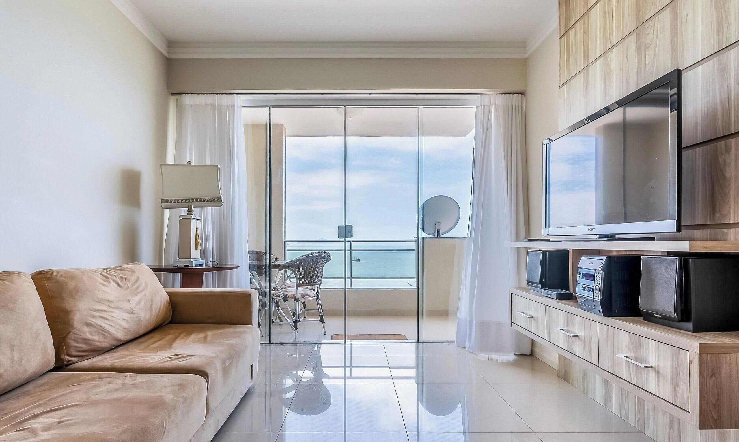 A modern living room with a sofa, a flat-screen TV, and a view of the sea through a glass door.