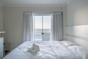 A hotel room with a bed, a small table, and a view of the sea through a large window. - Cozy 3BR apt W/balcony sea View Cet1702 (Balneário Camboriú)