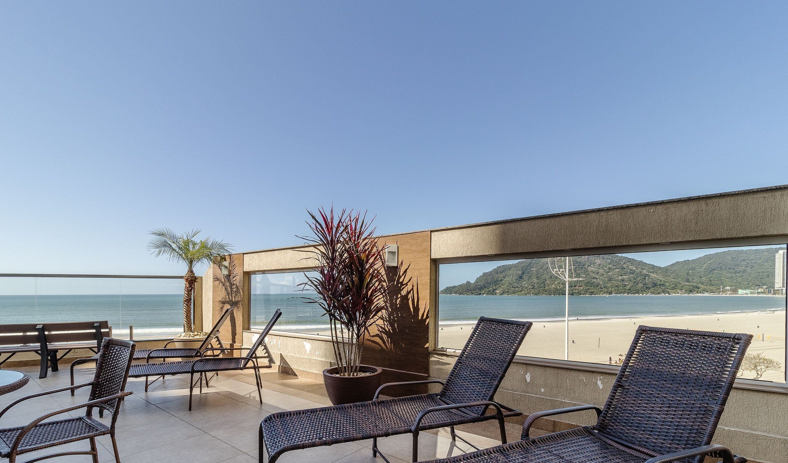 A terrace with wicker chairs and a potted plant, overlooking a beach and a mountain.