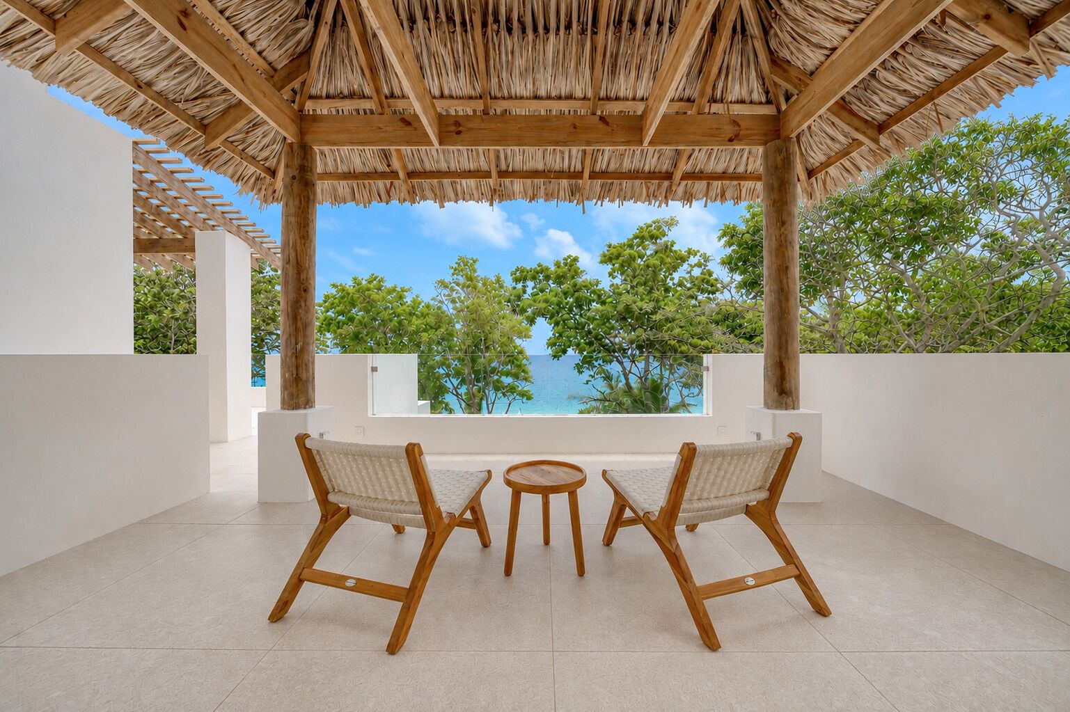 Un espace extérieur abrité, avec deux chaises en bois et une petite table, offrant une vue panoramique sur les arbres et l’océan.