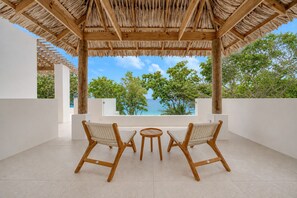 A covered outdoor seating area with two wooden chairs and a small table, overlooking a scenic view of trees and the ocean.