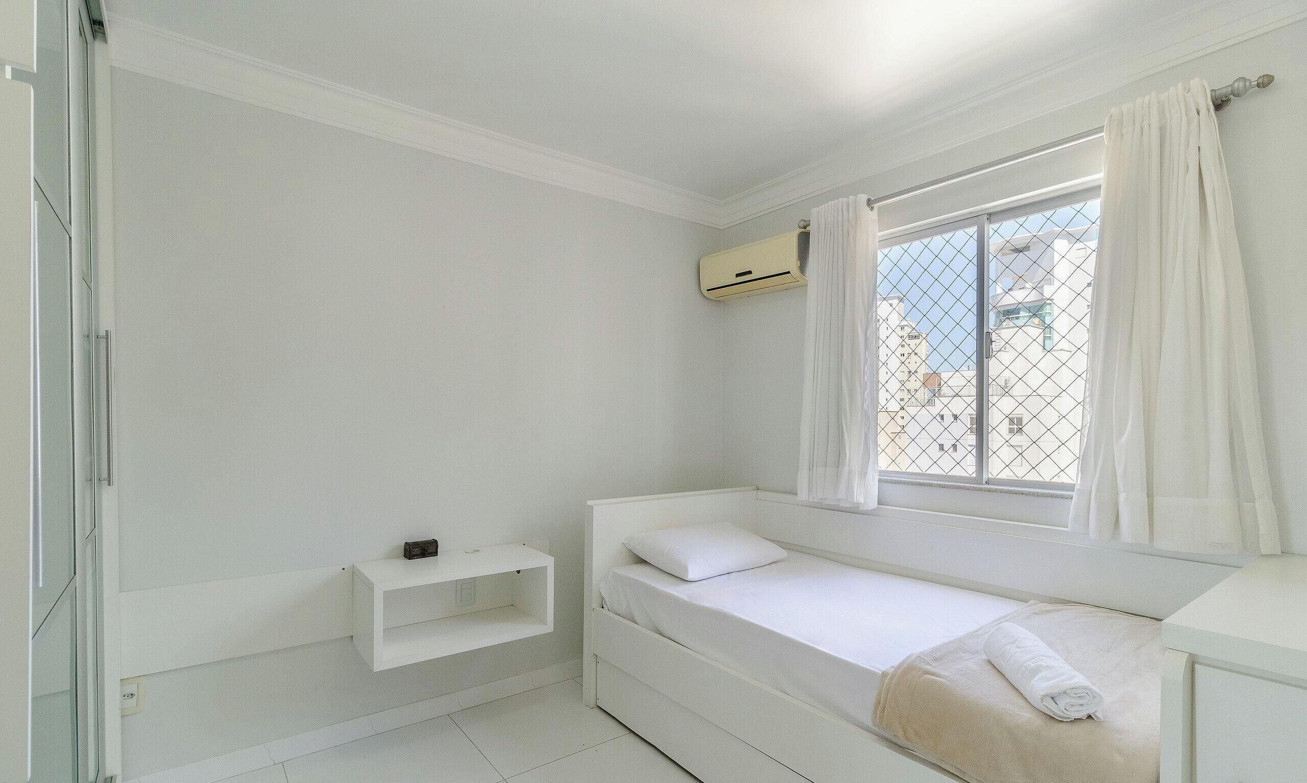 Habitació petita, de color blanc, amb un llit individual, una finestra, una taula de nit i un aparell d'aire condicionat.